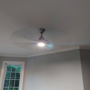 Dub Electric - Study - A ceiling fan with a built-in light fixture is installed on a plain white ceiling. The fan's light is turned on, emitting bright illumination. A window with white trim and multiple panes is visible on the left side, revealing green foliage outside. The room has light-colored walls and crown molding along the ceiling edge. - Electricians Near Me - Grapevine, Texas