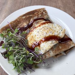 Bacon & Brussel Sprout Crepe - A savory crepe topped with a fried egg, drizzled with sauce, served with microgreens on a white plate.