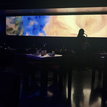 Vybe - Saxophone player - A person is playing the saxophone in a dark indoor setting, likely a restaurant or lounge, with a large, illuminated digital screen displaying abstract colors and patterns in the background. The venue has tables set with condiments and napkins, and the overall lighting is low, emphasizing the performer and the screen. - Venues & Event Spaces Near Me - Chandler, Arizona