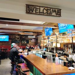 The image shows the interior of a bar or restaurant with a wooden bar counter and several patrons seated on bar stools. Behind the bar, there are shelves stocked with bottles of alcohol, and multiple TV screens mounted on the wall displaying content. The decor includes wooden beams and a rustic sign that says 'WELCOME' hanging above. The setting appears casual and lively, with some patrons engaged in conversation.