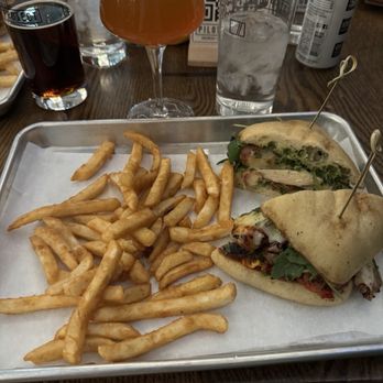 Pilot Project Brewing - Milwaukee - Chicken Pesto Melt - French fries and two sandwiches with skewers are arranged on a metal tray lined with parchment paper. The sandwiches contain ingredients like greens, cheese, and meats. In the background, there are various drinks including a glass of dark soda, a wine glass with a pinkish beverage, and a glass of water. The setting appears to be a casual dining restaurant with a wooden table and QR code signs. - Brewpubs Near Me - Milwaukee, Wisconsin