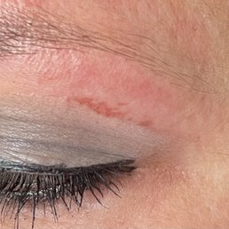 The image shows a close-up of a person's closed eye, highlighting the eyelid with makeup, including eyeliner and eyeshadow. The skin around the eye displays natural texture and a small scar or mark on the eyelid. Fine eyelashes are visible, and the skin tone appears even with some natural variations.