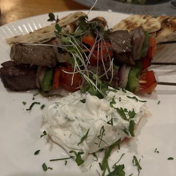 Molos - Skewered beef and vegetable kebabs, including pieces of beef, cherry tomatoes, and green peppers, are arranged on a white plate. A dollop of herbed cream cheese is placed beside the skewers, garnished with chopped herbs. Microgreens are used as a garnish on top of the skewers. The background shows a wooden table and another dish in a gray plate. - Greek Near Me - Weehawken, New Jersey