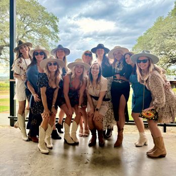 360 Transportation - A group of twelve women dressed in western-themed clothing, including cowboy hats, sunglasses, and boots, pose together on a covered outdoor patio with a scenic background of trees and cloudy sky. They are smiling and appear to be enjoying a social gathering or event. - Buses Near Me - Austin, Texas