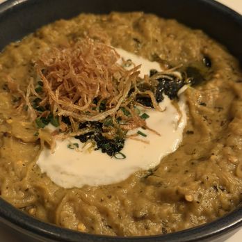 Joon - Eggplant - A close-up of a bowl containing a cooked eggplant dish, topped with fried onions, a dollop of cream, and chopped herbs. The dish appears to be a Persian or Middle Eastern style eggplant preparation, served in a black bowl. The background shows a blurred table setting, indicating a restaurant environment. - Persian/Iranian Near Me - Vienna, Virginia