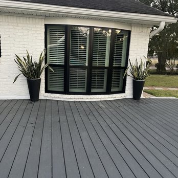 Upright Windows - The image shows the exterior of a house with white brick walls and a large bay window with black framing and white horizontal blinds. Two tall, black planters with green and yellow variegated plants are positioned on either side of the window. The house has a dark shingled roof with visible vents and a white gutter system. The gray wooden deck extends in front of the window, with a grassy yard and trees visible in the background. - Windows Installation Near Me - Klein, Texas