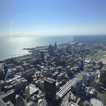 Skydeck Chicago - The image captures a cityscape of Chicago from a high vantage point, showing numerous tall buildings and skyscrapers in the downtown area. The layout includes streets and green spaces, with Lake Michigan visible on the horizon. The sky is clear and blue, indicating a sunny day. The photograph appears to be taken from a tall building or observation deck, providing a broad view of the city and lake. - Observatories Near Me - Chicago, Illinois