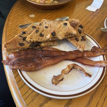 Bev's Cafe - Skillet and raisin toast and bacon - A breakfast plate contains two strips of cooked bacon, raisin toast slices, and a small piece of bacon on a wooden table. In the background, there is a bowl of hash browns with ham and cheese, along with a spoon and some paper napkins. The setting appears to be a casual dining environment. - American Near Me - Red Wing, Minnesota