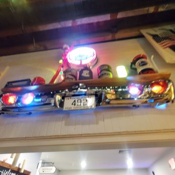 Station 101 - A wall-mounted decorative piece includes a chrome bumper, several cans, a license plate, and colorful lights, arranged on a wooden board. The setup is indoors, with a ceiling and other objects visible in the background, including a newspaper and an American flag. The lighting is dim, emphasizing the illuminated elements of the decoration. - Beer Bar Near Me - Milford, New Hampshire