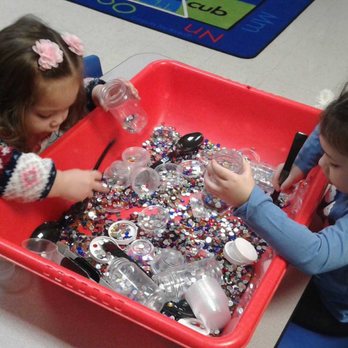 Wilkins Learning Center - Sensory play while learning! - Two young girls are seated at a table with a large red bin filled with colorful rhinestones, small containers, and tools. One girl is wearing a patterned sweater with flowers in her hair, while the other is dressed in a blue jacket with hair accessories. They are sorting or playing with the rhinestones, engaging in a sensory and fine motor activity in a classroom environment. - Preschools Near Me - Gilbert, Arizona