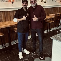 Two men are standing inside a restaurant with a mural of Middle Eastern landmarks on the wall behind them. One man is pointing at the other, and both are smiling.