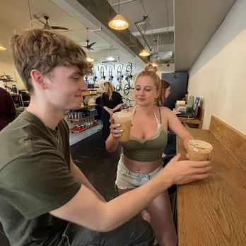 The Nitro Bar- Providence - A young man and woman are standing at a coffee shop counter, each holding a plastic cup of iced coffee. The woman is wearing a green crop top and shorts, while the man is dressed in a dark t-shirt. The background shows the coffee shop interior with a barista working behind the counter, and other customers present. The setting includes warm lighting and a casual atmosphere. - Coffee & Tea Near Me - Providence, Rhode Island