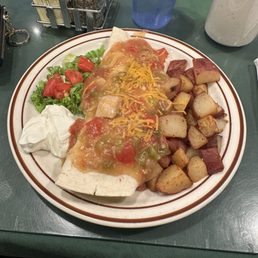 Breakfast Burrito - The image shows a breakfast plate with a large burrito topped with melted cheese and salsa, accompanied by crispy hash browns, a small side salad with chopped tomatoes and lettuce, and a serving of sour cream. The plate is set on a green table with condiments and utensils nearby, suggesting a casual dining setting.
