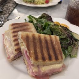 Ham & Gruyere Panini - A close-up of a ham and Gruyère cheese panini with toasted grill marks, cut into two halves, served alongside a mixed green salad with leafy lettuce. The meal is presented on a white plate, with a beverage and another salad visible in the background, on a marble table setting.