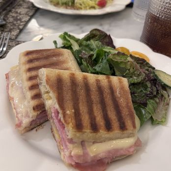 August Cafe - Ham & Gruyere Panini - A close-up of a ham and Gruyère cheese panini with toasted grill marks, cut into two halves, served alongside a mixed green salad with leafy lettuce. The meal is presented on a white plate, with a beverage and another salad visible in the background, on a marble table setting. - Cafes Near Me - New York, New York