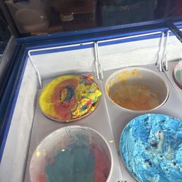 Superman Ice Cream, Blue Moon ice cream etc... - Four tubs of ice cream with vibrant colors are shown in a freezer display case, including yellow, blue, and mixed colors.