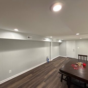 One Stop Handy - New drywall and finish where four air vents are, new paneling on the wall where the door is. - The image shows a spacious basement room with gray painted walls and wood flooring. The ceiling has recessed lighting fixtures, and a central ceiling light is turned on. There is a white door on the far wall, and a few items, including a water bottle, are placed on the floor near the wall. On the right side, a dark wooden dining table with chairs is visible, with some objects on the table, such as a red cup and other small items. The room appears clean and minimally furnished. - Handyman Near Me