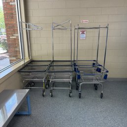 They have plenty of laundry carts that are clean & new! - Three metal shopping carts are positioned against a beige cinder block wall near a window in an indoor space, with a bench nearby.