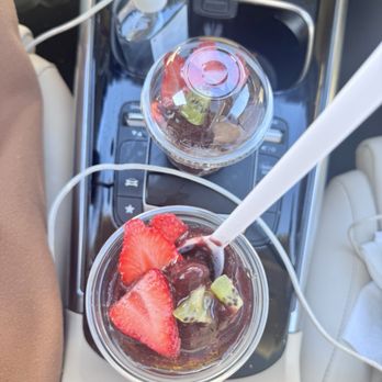 Deep Purpl - The image shows two clear plastic cups with fruit-topped desserts on a car's center console. One cup contains a dark-colored dessert with strawberries and kiwi slices, with a white straw inserted. The other cup has a lid and contains a dark beverage, possibly iced coffee or soda. The console also has a hand sanitizer bottle and a charging cable visible. - Juice Bars & Smoothies Near Me - Chicago, Illinois