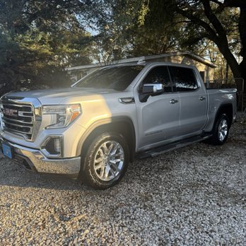Auto Reflections Window Tinting - A silver GMC Sierra pickup truck with four doors and tinted windows is parked on a gravel surface outdoors. The truck's front grille and headlights are visible, and the vehicle appears clean and well-maintained. Surrounding the truck are trees with sunlight filtering through the branches, creating a natural setting. The truck is positioned at an angle, showing both the front and side views, with a background of a house and foliage. - Car Window Tinting Near Me - Spring, Texas