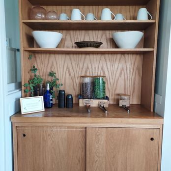Willowbrook Spa - Treats - A wooden shelving unit holds white ceramic cups, bowls, and small containers, with some labeled jars containing herbs or ingredients, and a small potted plant. The setting appears to be part of a wellness spa or herbal treatment area, with a clean and organized presentation of items used for treatments or services. - Estheticians Near Me - Canandaigua, New York