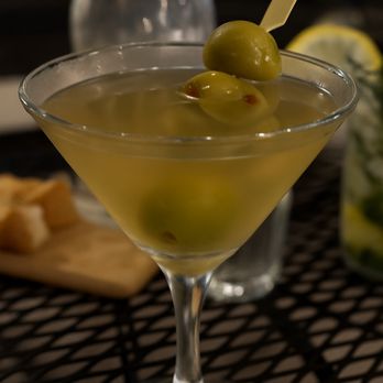 Sunset Grill - dirty martini - A cocktail served in a martini glass, garnished with green olives on a pick, with a blurred background including a water bottle and lemon slices. - Steakhouses Near Me - Moab, Utah