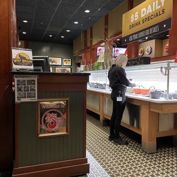 Ruby Tuesday - Ruby Tuesday staff attending to the Garden Bar - A woman with blonde hair, wearing a black uniform, is preparing food or handling items behind the counter at Ruby Tuesday in Amsterdam, NY. The counter area features a glass display case, and there are signs and decorations on the wall, including a large yellow sign advertising $5 daily drink specials. The floor has a patterned tile design, and the restaurant interior includes framed artwork and plants. - American Near Me - Amsterdam, New York