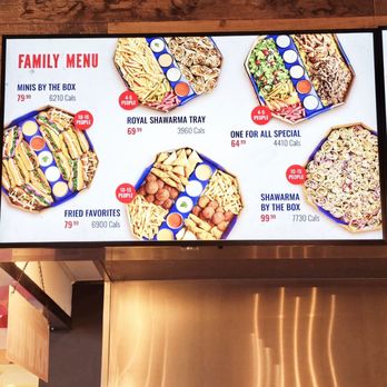 Tahini's - Family menu - The image features a digital menu board displaying family meal options at a restaurant, including items like Minions by the Box, Royal Shawarma Tray, One for All Special, Fried Favorites, and Shawarma by the Box. Each option lists the price, calorie count, and serving size, with some marked for specific group sizes. The menu is set against a wooden background with a stainless steel structure below. - Mediterranean Near Me - Pickering, Ontario