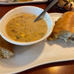 Steak and Cheddar - A white rectangular plate holds a small bowl of corn chowder with visible corn kernels and herbs. Next to the bowl are slices of plain bread and a bread roll with a dark filling inside, possibly a sweet or savory ingredient. The plate is on a wooden table, with part of another dish visible in the background.