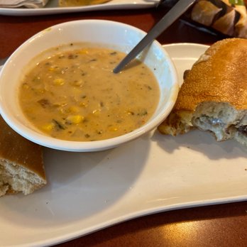 Panera Bread - Corn chowder, steak and cheddar - A white rectangular plate holds a small bowl of corn chowder with visible corn kernels and herbs. Next to the bowl are slices of plain bread and a bread roll with a dark filling inside, possibly a sweet or savory ingredient. The plate is on a wooden table, with part of another dish visible in the background. - Salad Near Me - Branson, Missouri