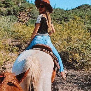 Western Destinations - A woman wearing a wide-brimmed hat, sunglasses, and casual clothing rides a white horse through a desert area with cacti and bushes. - Venues & Event Spaces Near Me - Phoenix, Arizona