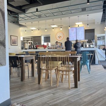 Epic Coffee - The image shows the interior of a coffee shop with a modern design, including a counter with digital menu screens and a logo. Customers are standing at the counter, and there are various chairs and tables for seating. The ceiling has exposed ductwork and hanging light bulbs, and the floor is wood with a patterned rug in the foreground. - Juice Bars & Smoothies Near Me - Carmel, Indiana
