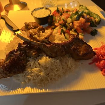 Selda - Lamb chops - A plate with lamb chops, rice, a fresh salad, and a small container of sauce, set on a table with a glass and a lamp in the background. - Mediterranean Near Me - Irving, Texas