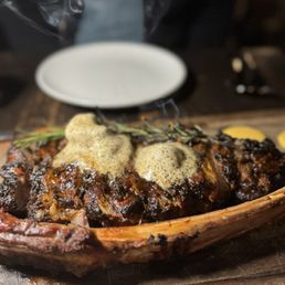A steak dish with herb and peppercorn sauce is presented on a wooden platter, with a sprig of herbs and a small bowl of sauce nearby.