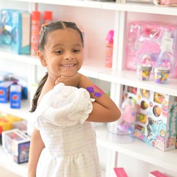 Buzz Cuts Kids Salon - A happy girl poses in front of a shelf filled with salon products and fun toys. Her arm has a purple glittery design, and she looks ready fo - Kids Hair Salons Near Me - Plano, Texas Buzz Cuts Kids Salon - A happy girl poses in front of a shelf filled with salon products and fun toys. Her arm has a purple glittery design, and she looks ready fo - Kids Hair Salons Near Me - Plano, Texas