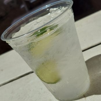 Ideal Bar - a drink in a plastic cup - A clear plastic cup filled with a fizzy, lemon-lime flavored beverage, with visible bubbles and lime slices floating inside. The cup is placed on a light-colored surface, and condensation can be seen on the outside of the cup, indicating the drink is cold. - Dive Bars Near Me - Madison, Wisconsin