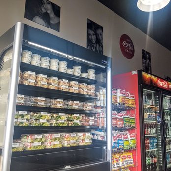 Shelly's Kitchen - a display of food in a store - The image shows a refrigerated display case with a glass front, filled with pre-packaged salads, sandwiches, and baked goods. To the right, there is a vending machine with snacks and beverages, including soda and bottled drinks. The setting appears to be a convenience store or similar retail environment, with posters and photographs on the wall above the display. - Pizza Near Me - Plaistow, New Hampshire