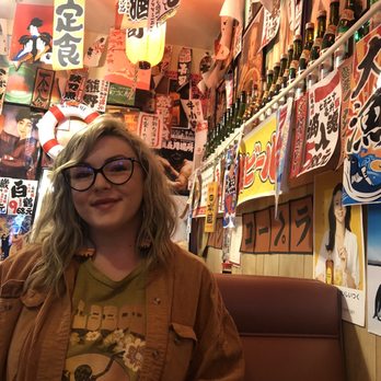 Dragon Sushi - My beautiful date. - A woman with glasses and wavy blonde hair is seated in a Japanese-themed restaurant, surrounded by colorful posters, banners, and bottles on the wall. The decor includes Japanese characters, images, and traditional elements, creating an authentic atmosphere. She is wearing a brown jacket over a graphic T-shirt and is smiling at the camera. A cup of tea or coffee is visible in the foreground, and the restaurant's interior is densely decorated with various Japanese signs and artwork. - Sushi Bars Near Me - Albuquerque, New Mexico