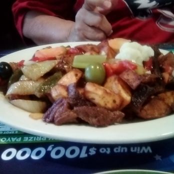 Baliza - My friend enjoyed her meal!! - A large oval white plate holds a serving of grilled vegetables, including zucchini, onions, and peppers, along with pieces of grilled meat. There are also mashed potatoes and black olives on the plate. The dish is placed on a table with a menu underneath, and a person wearing a red shirt is visible in the background, holding a utensil. - Portuguese Near Me - Kearny, New Jersey