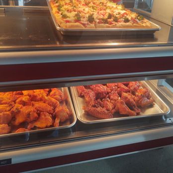 Shelly's Kitchen - Pizza & Wings - The image features a food display case with a tray of pizza on the top shelf, topped with vegetables and cheese. Inside the case, there are trays of fried chicken and chicken wings, arranged side by side. The case is made of metal and glass, with a red and white exterior, typical of casual eateries offering pizza and fried foods. - Pizza Near Me - Plaistow, New Hampshire