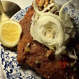 Pork Schnitzel - A plate contains a breaded and fried fish fillet, accompanied by a lemon wedge on the side. The fillet is topped with shredded onions, sliced peppers, and chopped nuts or seeds. The dish is served on a blue and white patterned ceramic plate, with other dishes partially visible in the background, indicating a meal setting.