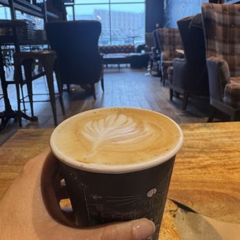 Ground Central - A person holds a black paper cup filled with a latte featuring a leaf-shaped latte art design. The cafe interior includes various chairs, a large window, and ambient lighting, creating a cozy atmosphere. The wooden table and background seating arrangements are visible, indicating a relaxed coffee shop environment. - Coffee & Tea Near Me - Lynbrook, New York