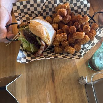 Wildfangs Grill House - A burger with bacon, lettuce, and a bun is presented on checkered paper, accompanied by a large serving of fried tater tots. The burger appears to have additional toppings and is held by a person. The setting is a casual dining environment with a wooden table, a glass of water, and a small sign visible nearby. - Barbeque Near Me - Springfield, Oregon