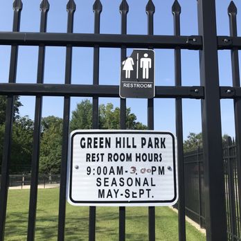 Green Hill Park - A black metal fence features a restroom sign and a park hours sign at Green Hill Park, with trees and a grassy area in the background. - Parks Near Me - Worcester, Massachusetts