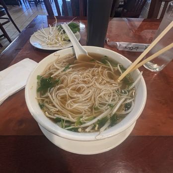 PHO 52 Frederick - Small Brisket Pho - A large white bowl filled with noodle soup containing broth, thin noodles, green onions, and bean sprouts is placed on a wooden table. A pair of chopsticks and a spoon rest on the bowl. Nearby, there is a glass of water, a black beverage container, a napkin, and a small plate with bean sprouts and lime slices. The setting appears to be a casual Vietnamese restaurant with wooden chairs and tables. - Vietnamese Near Me - Frederick, Maryland