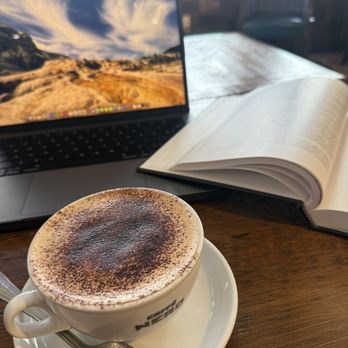 Caffè Nero - Tiramisu Latte - A coffee cup with a frothy drink, a laptop displaying a landscape, and an open book are arranged on a wooden table. - Coffee & Tea Near Me - Providence, Rhode Island