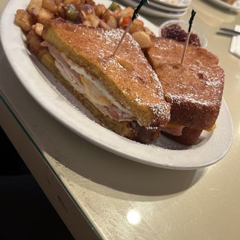 Mo's Egg House - Monte Cristo - A white oval plate holds a Monte Cristo sandwich, cut in half, with powdered sugar sprinkled on top. The sandwich contains ham, turkey, and cheese between slices of fried bread. Next to the sandwich are crispy hash browns with diced vegetables. Small containers of jam are also on the plate. The background shows a table with stacked plates, utensils, and additional food items, indicating a casual dining setting. - Breakfast & Brunch Near Me - Temecula, California