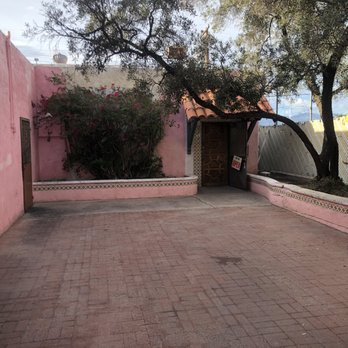 El Torero - An outdoor courtyard features a pink painted wall with two small windows and a wooden door with an 'OPEN' sign. There are bushes near the wall and a large tree with sprawling branches providing shade. The ground is paved with bricks, and a low pink wall borders the area. A chain-link fence is visible in the background, along with some structures and a cloudy sky. - Mexican Near Me - Tucson, Arizona