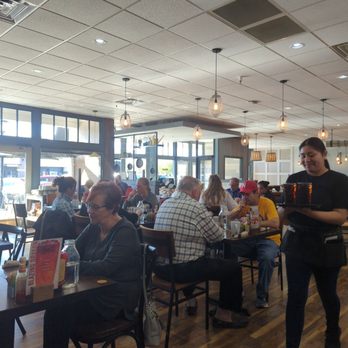 First Watch - Full house with great service! - The image shows a restaurant interior with multiple tables occupied by diverse customers. A server in black attire is carrying a tray with drinks, walking through the dining area. The restaurant has a modern design with pendant lighting, large windows, and wooden flooring. The atmosphere appears lively and well-lit, with natural light coming through the windows and ceiling lights illuminating the space. - Breakfast & Brunch Near Me - Mequon, Wisconsin