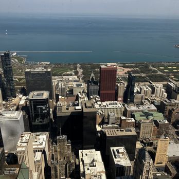 Skydeck Chicago - The image captures a cityscape of downtown Chicago from a high vantage point, featuring a dense cluster of tall buildings and skyscrapers. The foreground includes various commercial and office buildings, while the background reveals Lake Michigan with a shoreline, green spaces, and a few structures along the water. The scene emphasizes the urban density and proximity to the lake. - Observatories Near Me - Chicago, Illinois